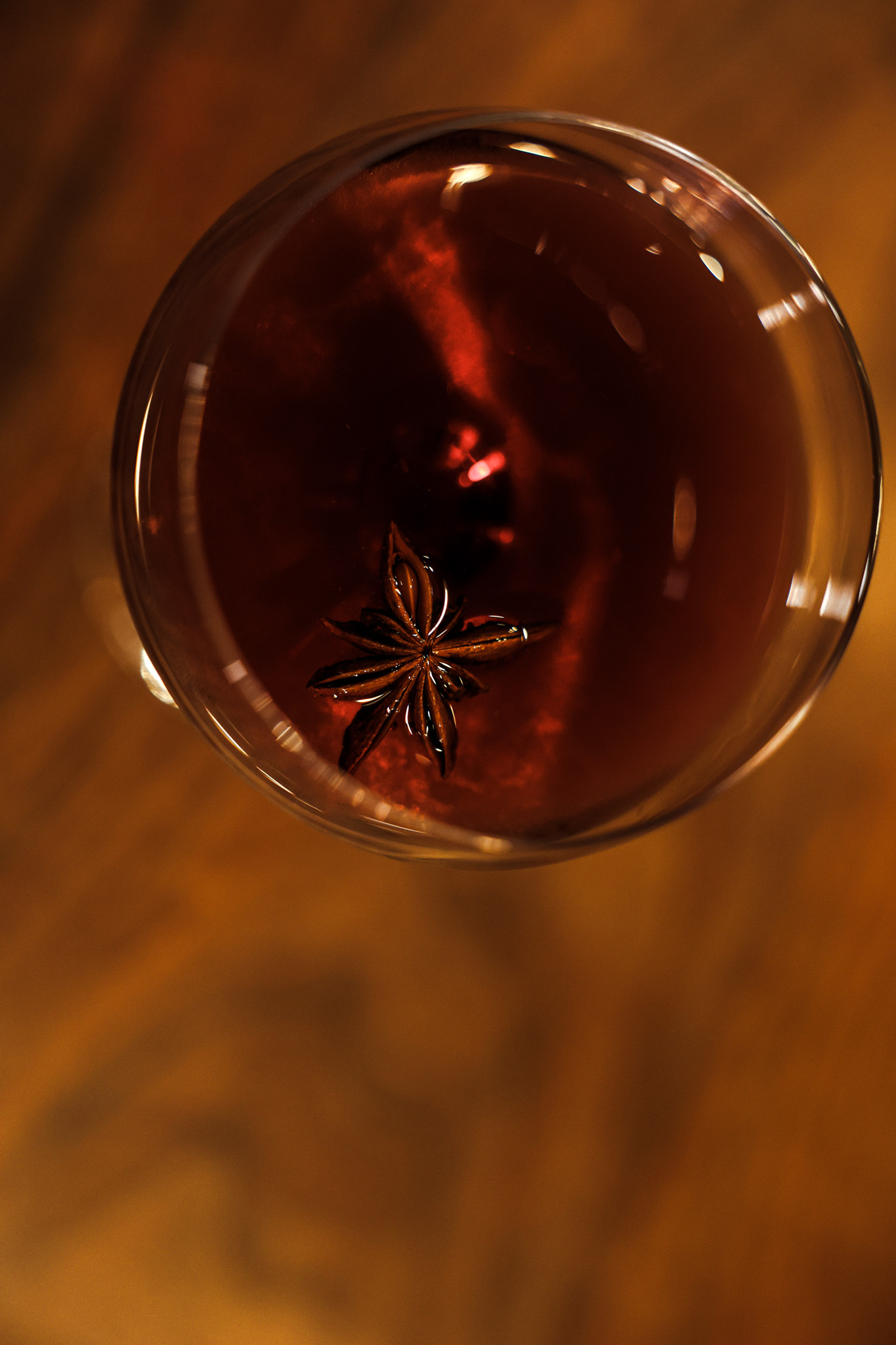 A dark cocktail in a coupe glass, photographed from above, with a star anise floating in oxblood-colored liquid against warm walnut.