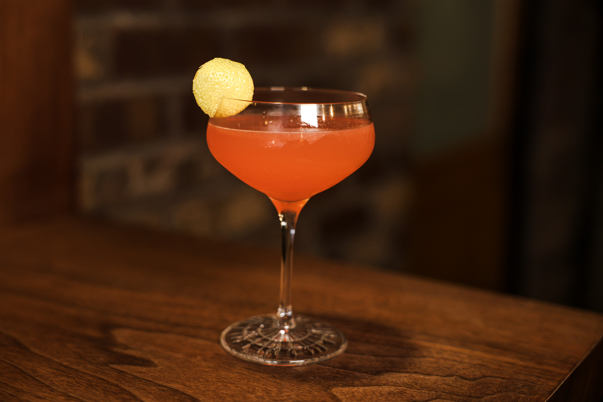 A coupe with citrus garnish on a walnut bar top, exposed brick wall blurred behind.