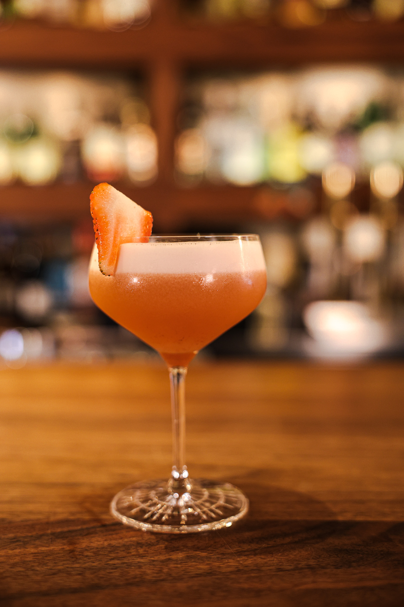 A coupe of an orange cocktail with a strawberry garnish on the bar, warm bottle wall in the background.